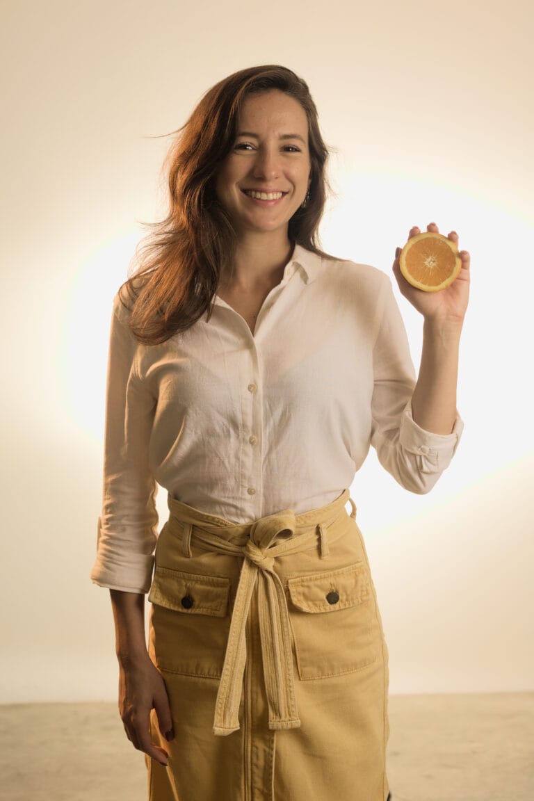 Virgínia Gaissionok, nutricionista, sorri enquanto segura metade de uma laranja em um ambiente bem iluminado.