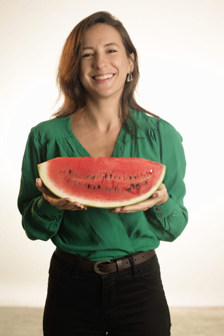 Virgínia Gaissionok sorrindo, segurando uma metade de melancia cortada com as mãos em um ambiente claro.