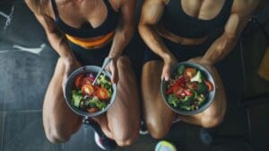 Duas mulheres fitness, vestindo roupas esportivas, estão sentadas lado a lado em um ambiente de academia, segurando tigelas de salada fresca e colorida. As tigelas contêm uma variedade de vegetais saudáveis, como brócolis, tomates, pimentões e folhas verdes. A imagem ilustra a importância da nutrição balanceada para mulheres atletas, destacando a relação entre alimentação saudável e desempenho físico. Palavras-chave: nutrição esportiva, alimentação saudável, dieta equilibrada, mulheres atletas, vegetais frescos, estilo de vida fitness.